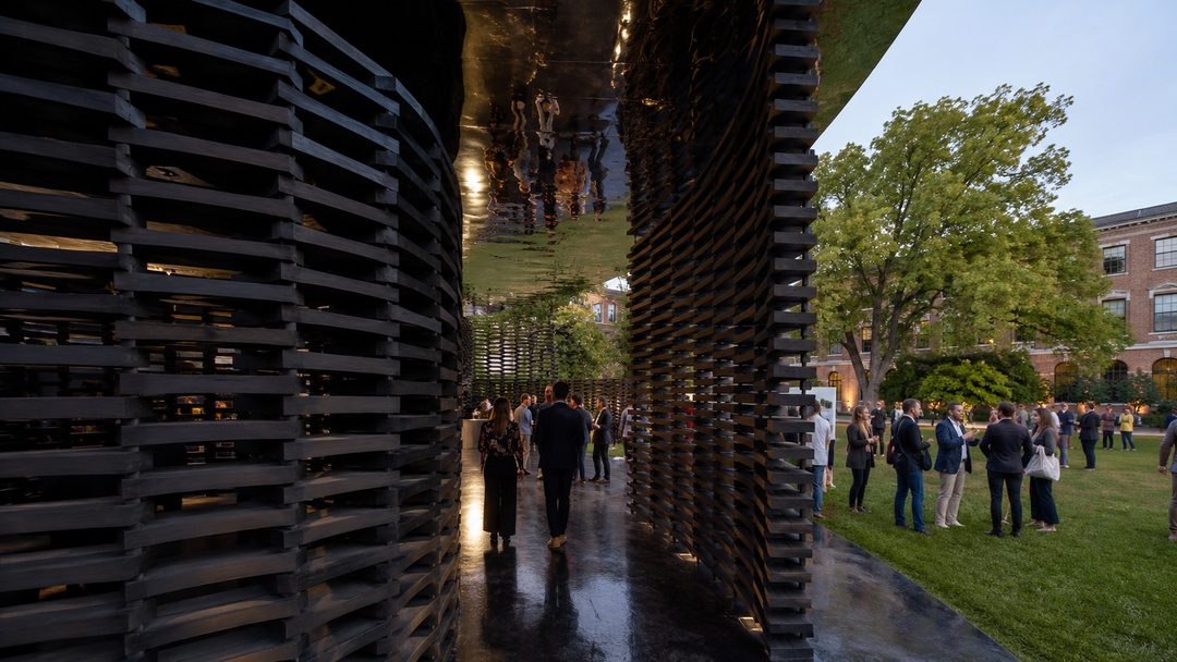 Pabell&oacute;n de arquitectura ef&iacute;mera con muros de listones negros, techo reflectante y visitantes recorriendo un jard&iacute;n exterior.