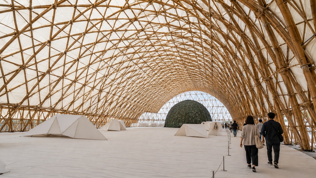 La imagen muestra el interior de una gran instalaci&oacute;n arquitect&oacute;nica temporal o pabell&oacute;n expositivo. El espacio est&aacute; cubierto por una estructura curva formada por ca&ntilde;as o elementos naturales entrelazados en una trama geom&eacute;trica repetitiva. La cubierta genera una b&oacute;veda amplia, luminosa y envolvente. En el interior se ven peque&ntilde;as estructuras blancas de forma triangular, un gran volumen esf&eacute;rico oscuro al fondo y varios visitantes recorriendo el espacio. La escena destaca la escala, la materialidad liviana, el car&aacute;cter inmersivo y la experiencia espacial propia de una arquitectura ef&iacute;mera y expositiva.