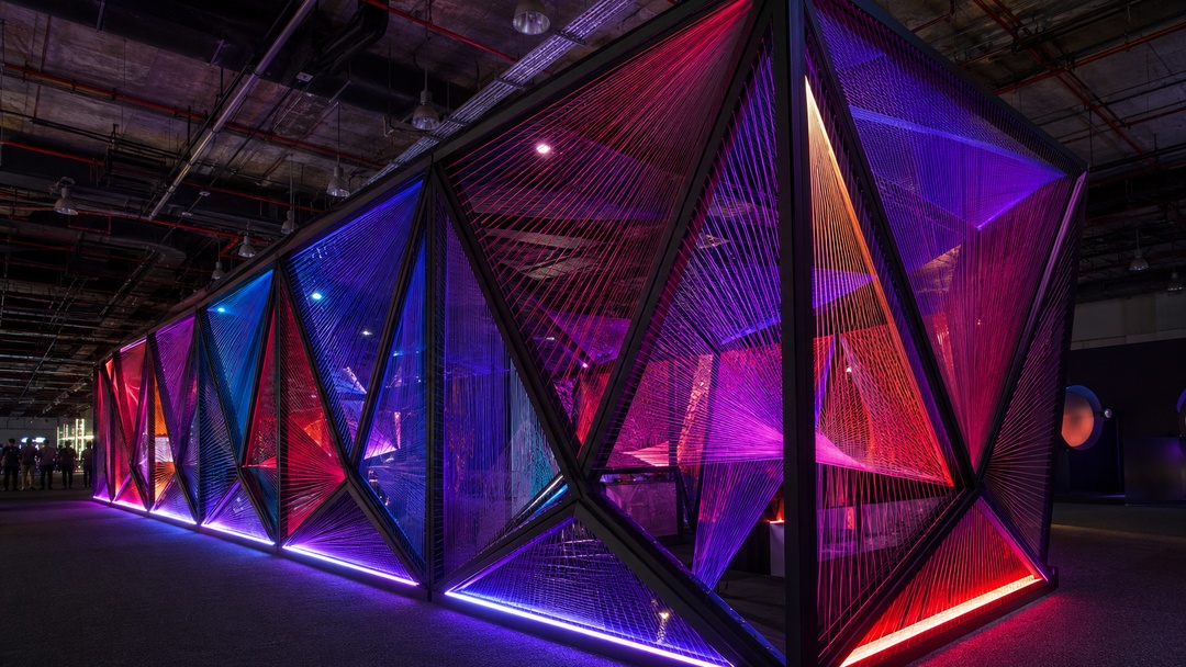 La imagen muestra una instalaci&oacute;n arquitect&oacute;nica temporal en un espacio interior oscuro, posiblemente un pabell&oacute;n de feria o exposici&oacute;n. La estructura est&aacute; formada por una sucesi&oacute;n de marcos triangulares negros, atravesados por hilos o tensores de colores que generan superficies geom&eacute;tricas transl&uacute;cidas. La iluminaci&oacute;n en tonos violeta, azul, rojo y naranja recorre la base y el interior de los m&oacute;dulos, creando una atm&oacute;sfera inmersiva, tecnol&oacute;gica y escenogr&aacute;fica. Al fondo se ven algunas personas, lo que permite dimensionar la escala recorrible de la instalaci&oacute;n.